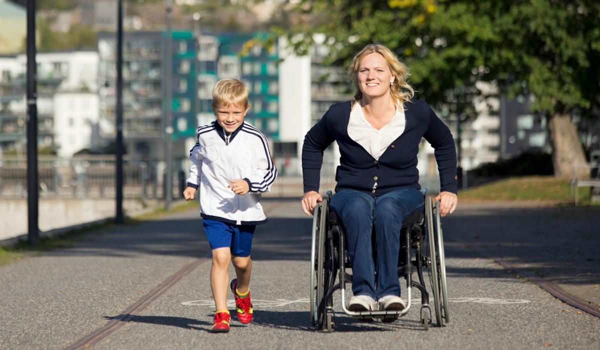 Foto på en kvinna som kör en rullstol. Bredvid henne springer ett barn. 