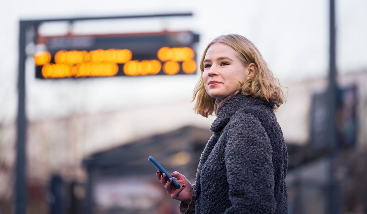 En kvinna står framför en busshållplats med en mobiltelefon i handen.