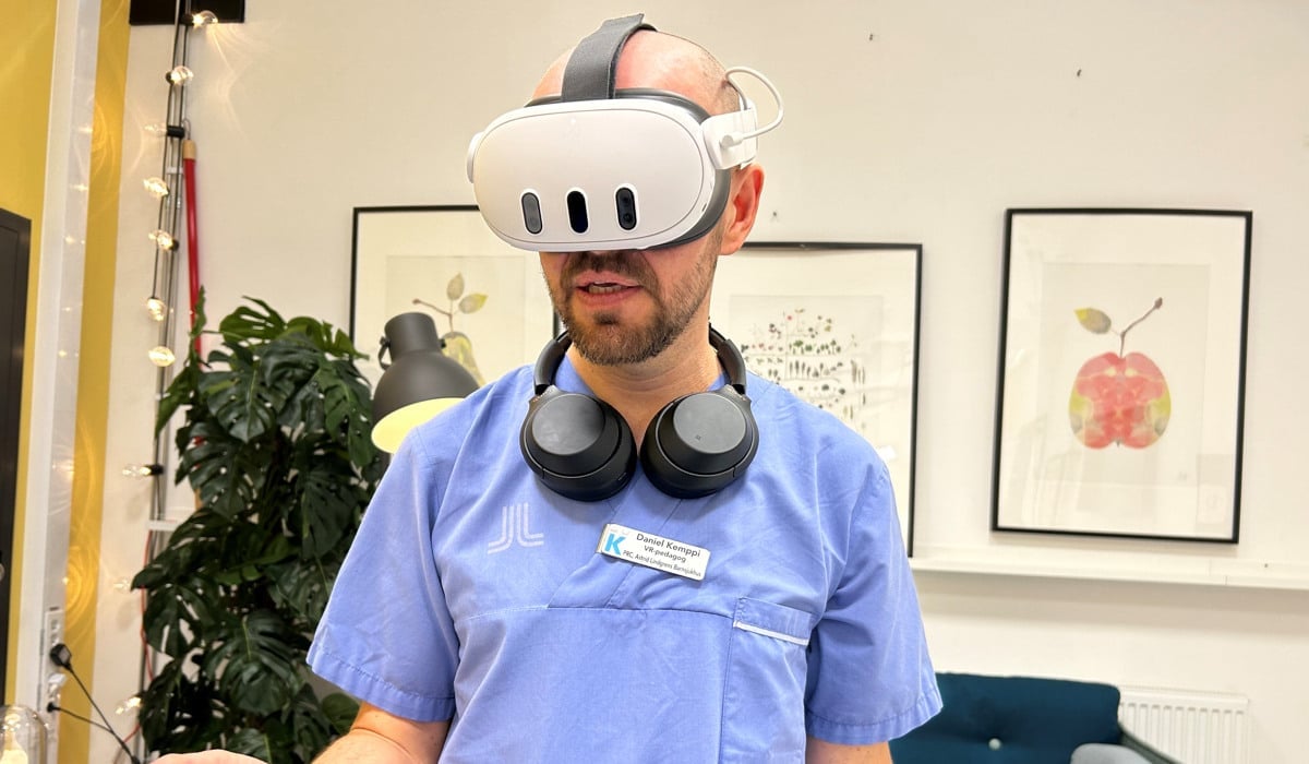 En man med blå sjukhusskjorta står med vr-headset på huvudet och handkontroller i handen