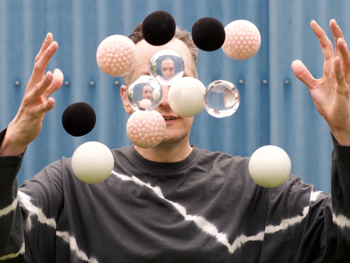 En person jonglerar med bollar. Framför ansiktet flyger svarta, vita och genomskinliga bollar.