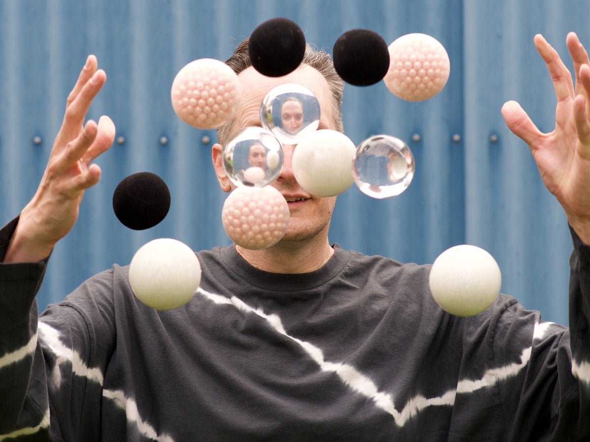 En person jonglerar med bollar. Framför ansiktet flyger svarta, vita och genomskinliga bollar.