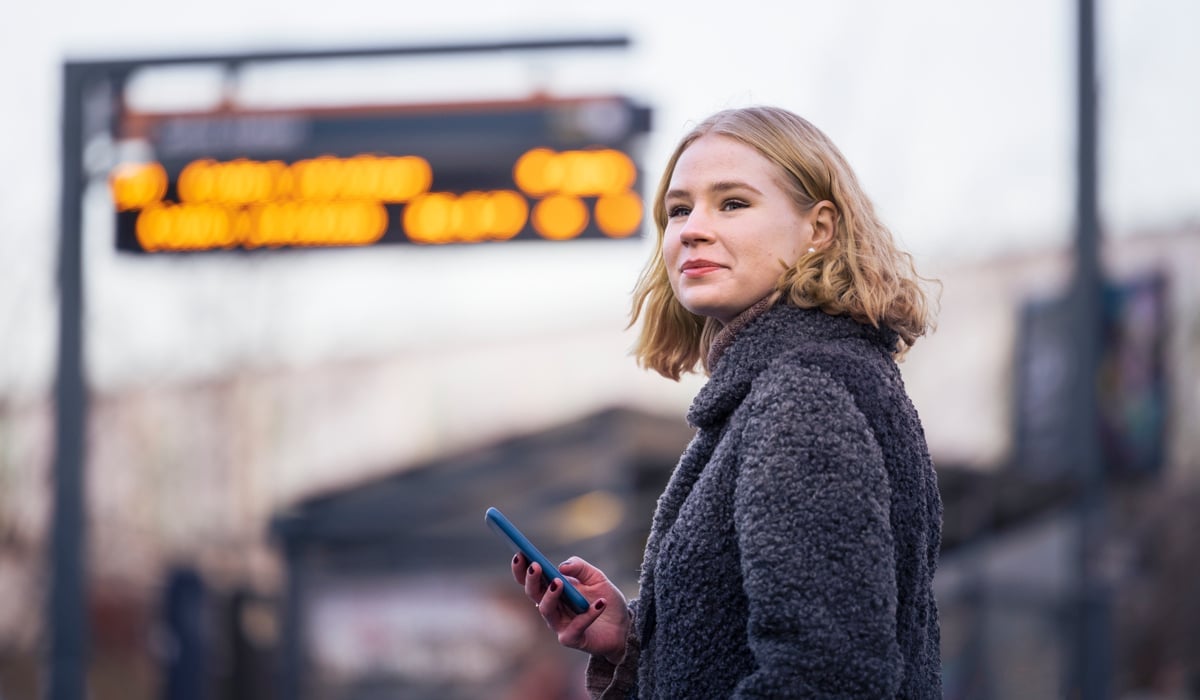 En kvinna står framför en busshållplats med en mobiltelefon i handen.