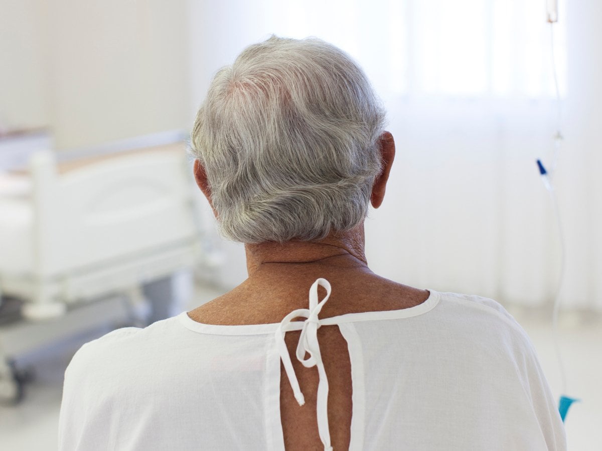 Äldre man i sjukhuskläder sitter i sjukhusmiljö med ryggen mot kameran