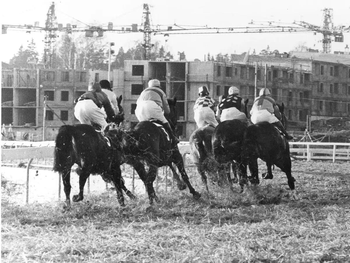 Svartvit bild av en klunga med hästar och ryttare. I bakgrunden byggs ett bostadsområde.