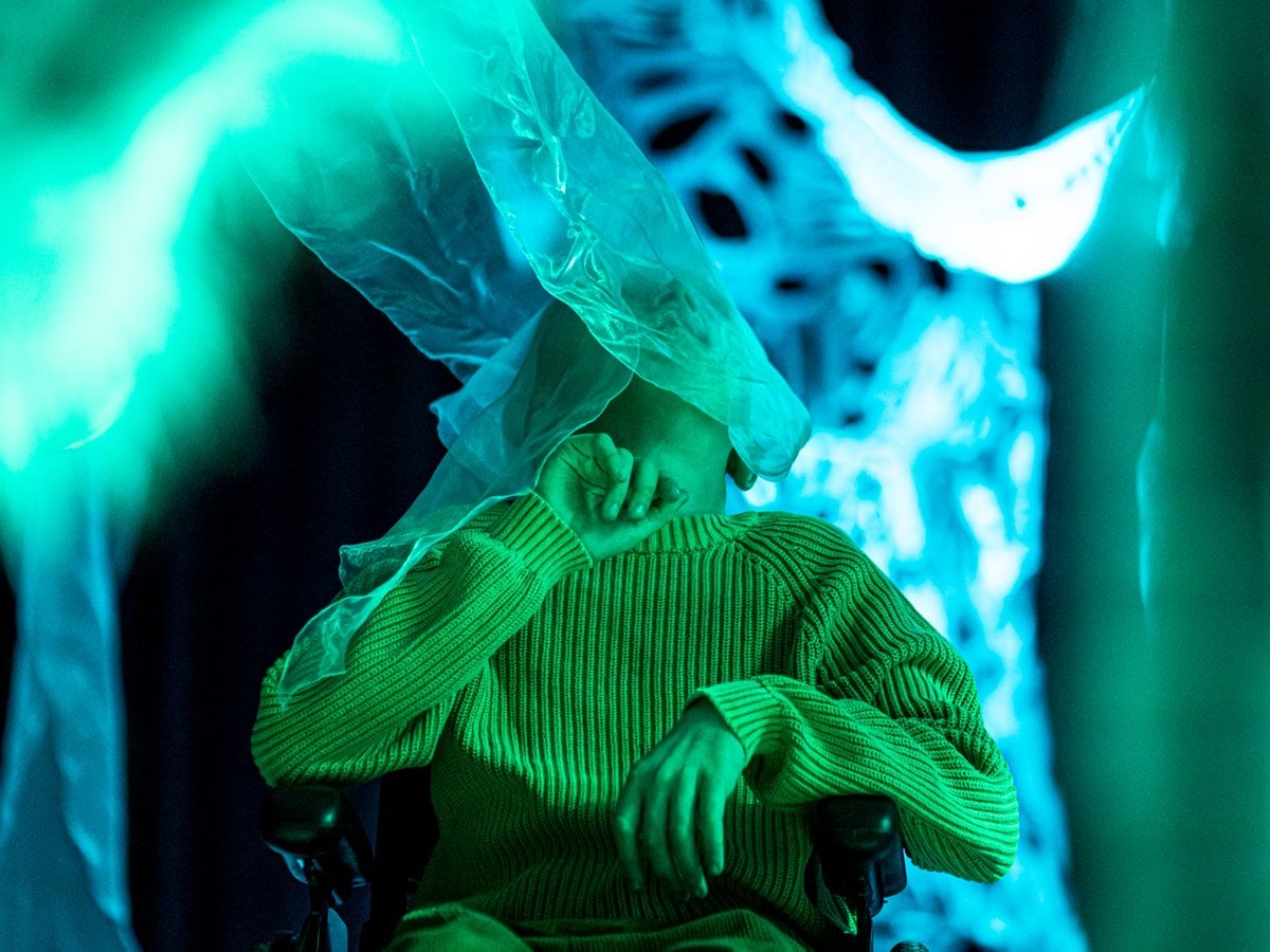 Barn i rullstol mitt i en upplevelseföreställning. Tunt tyg böljar över ansiktet.