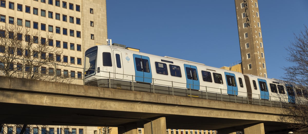 Tunnelbanetåg av modell C30 färdas över en bro. Blå himmel och kontorsbyggnad i bakgrunden.