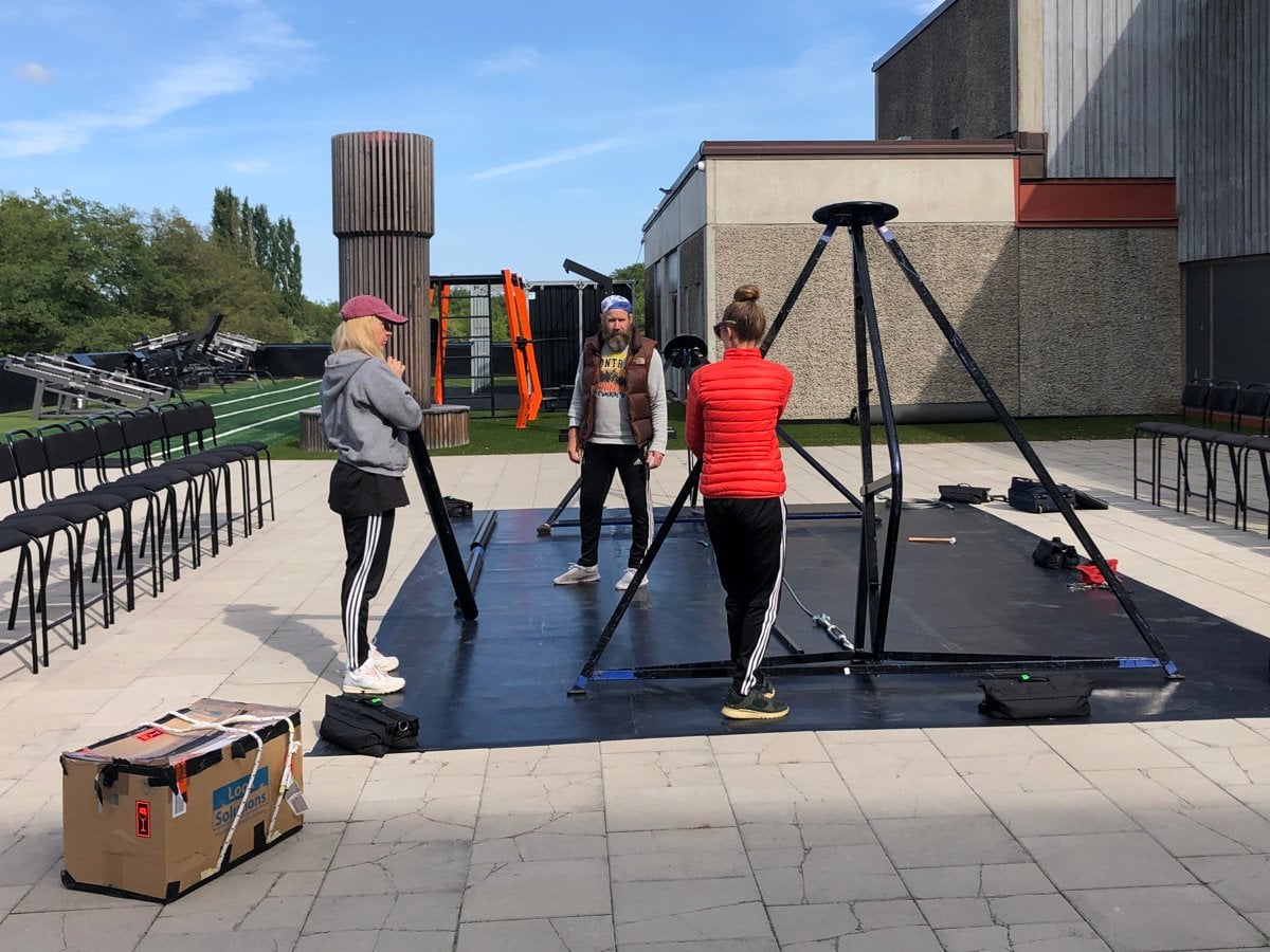 Tre personer i träningskläder står vid vid en ställning framför en byggnad av betong.