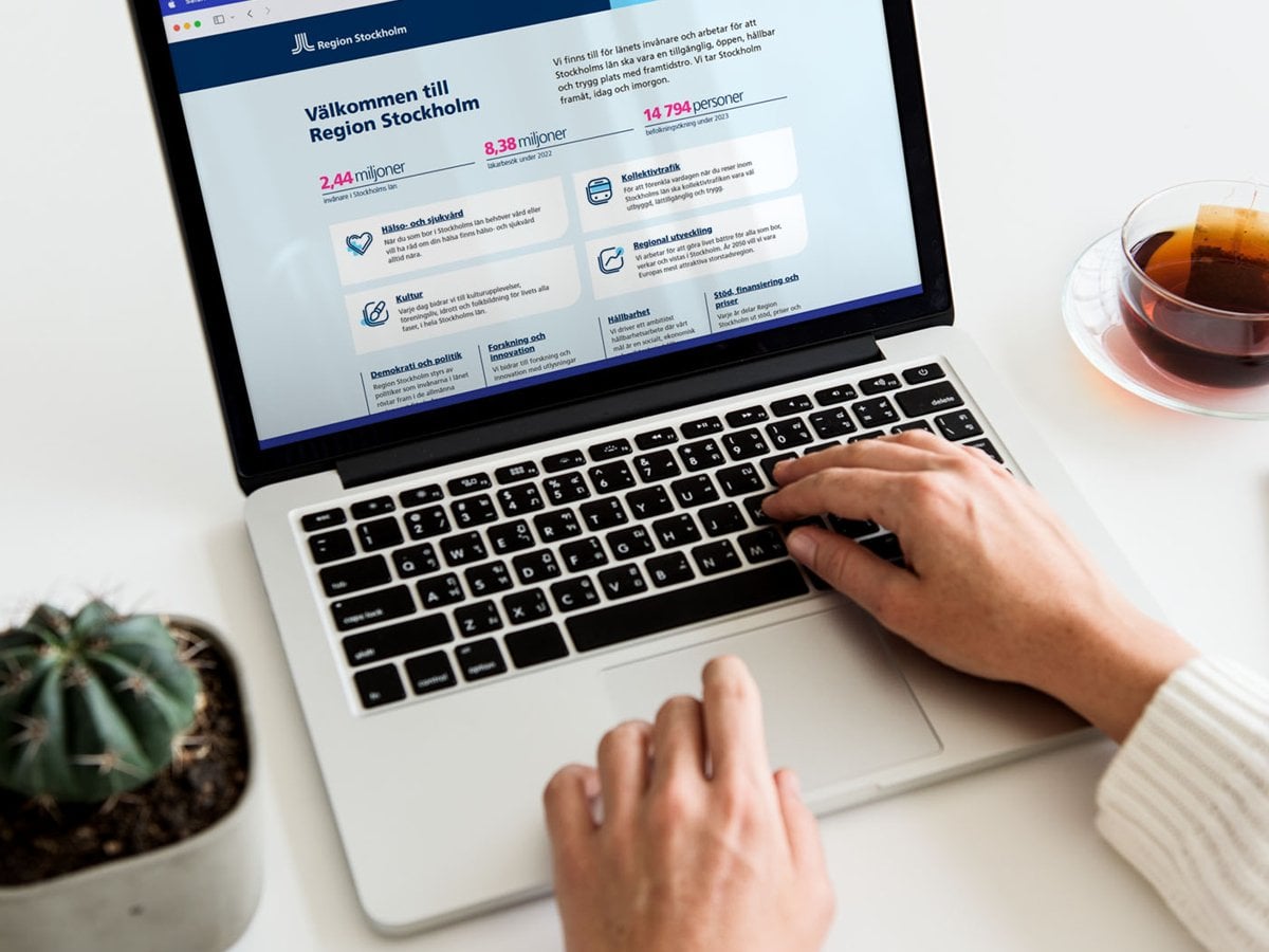 Foto på en person som använder en dator och har en mobiltelefon bredvid datorn. På datorn och mobiltelefon är Region Stockholms webbsida öppen