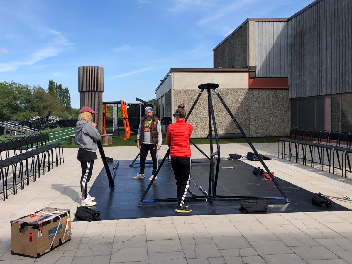 Tre personer i träningskläder står vid vid en ställning framför en byggnad av betong.