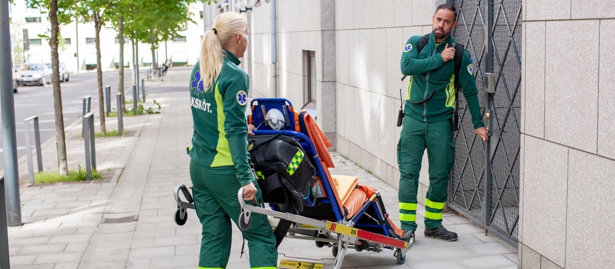 Två medarbetare i gröna arbetskläder väntar på att bli insläppa genom en grind. Den kvinnliga medarbetaren håller i en bår