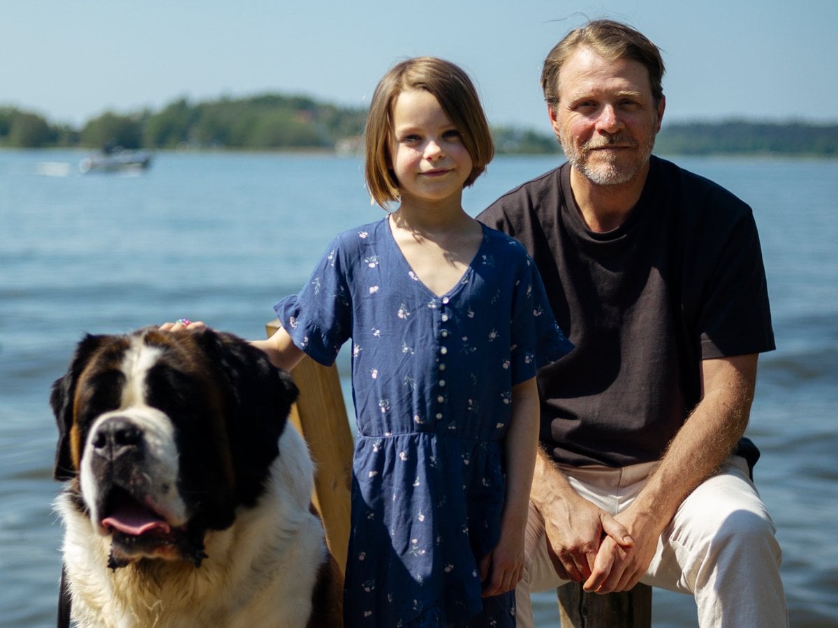 En stor hund, en flicka och hennes pappa tittar in i kameran i skärgårdsmiljö.