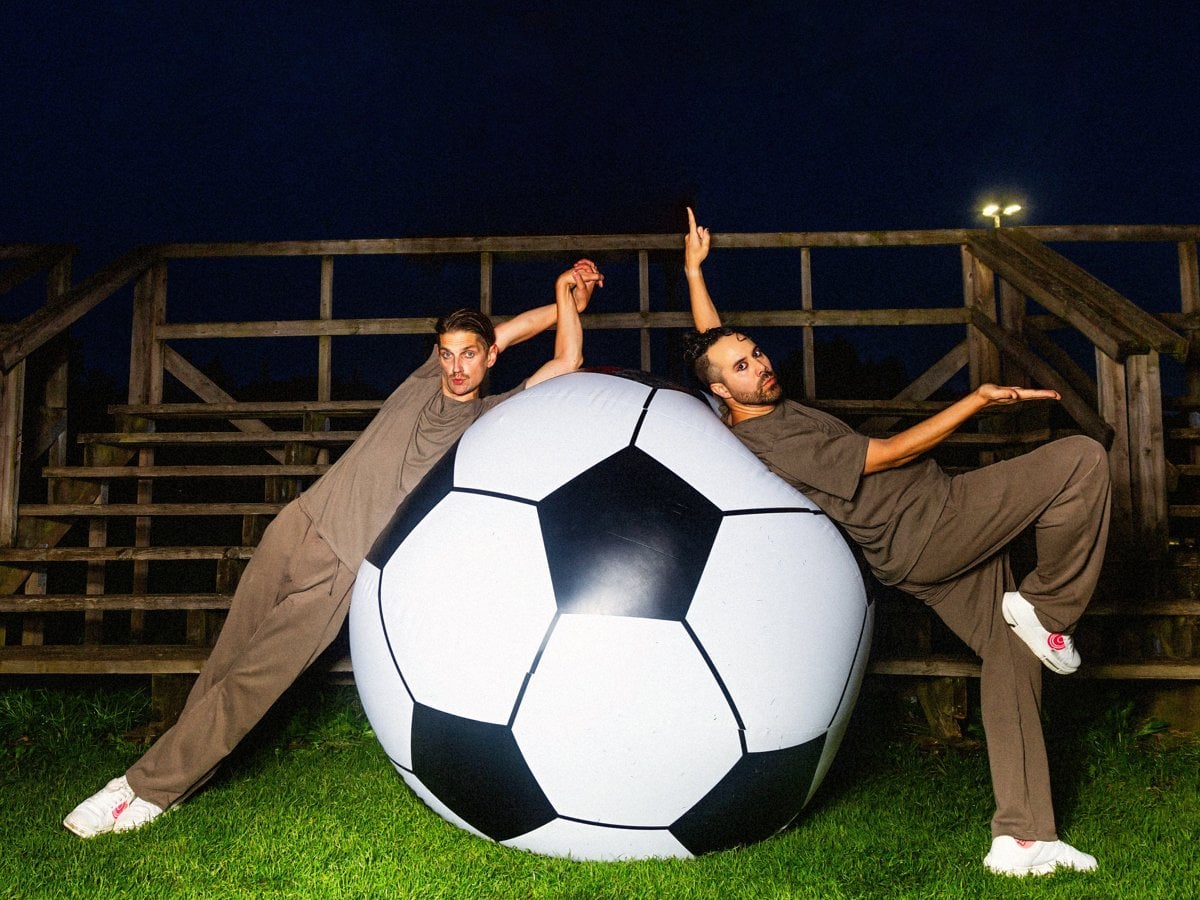 Två män i bruna träningsoveraller och vita sneakers lutar sig elegant mot en jättestor fotboll på en konstgräsplan framför en läktare på natten.