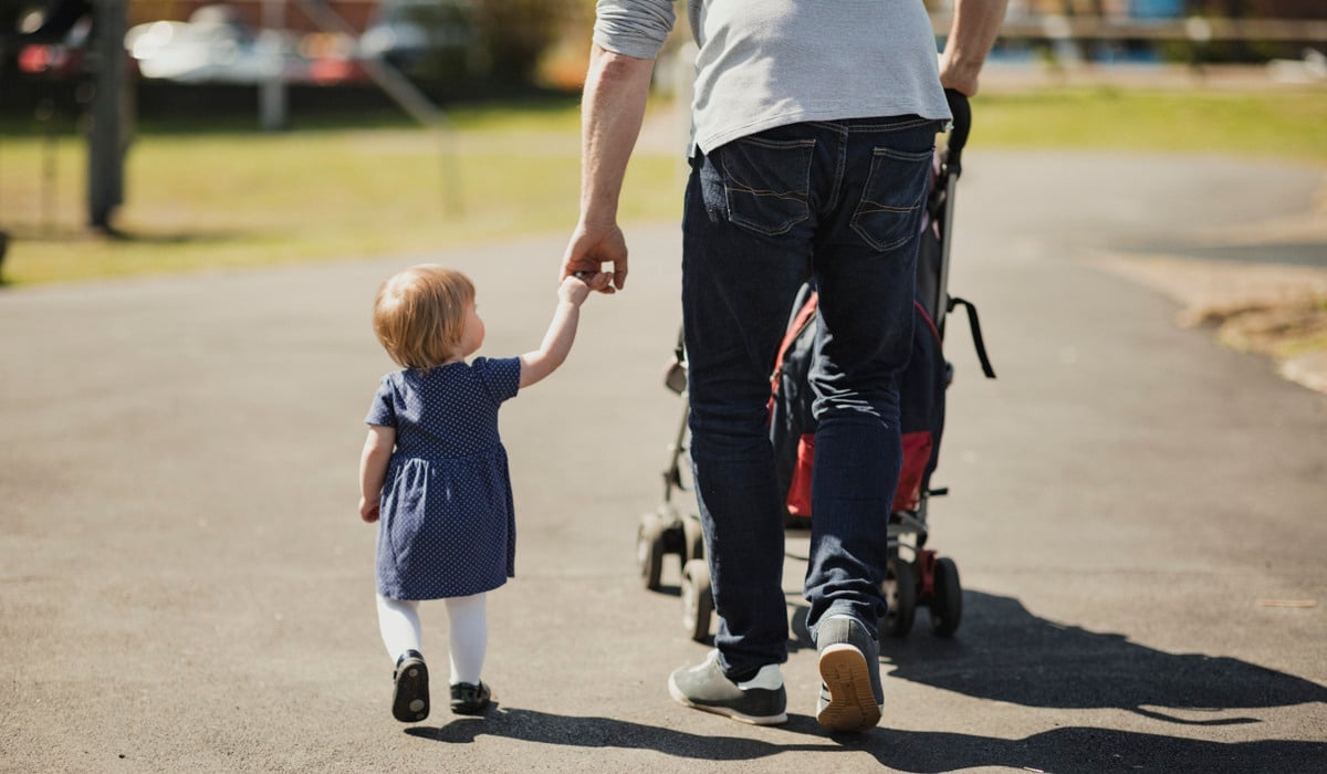 Liten flicka på promenad med sin pappa. Hon håller honom i handen och han i en barnvagn.
