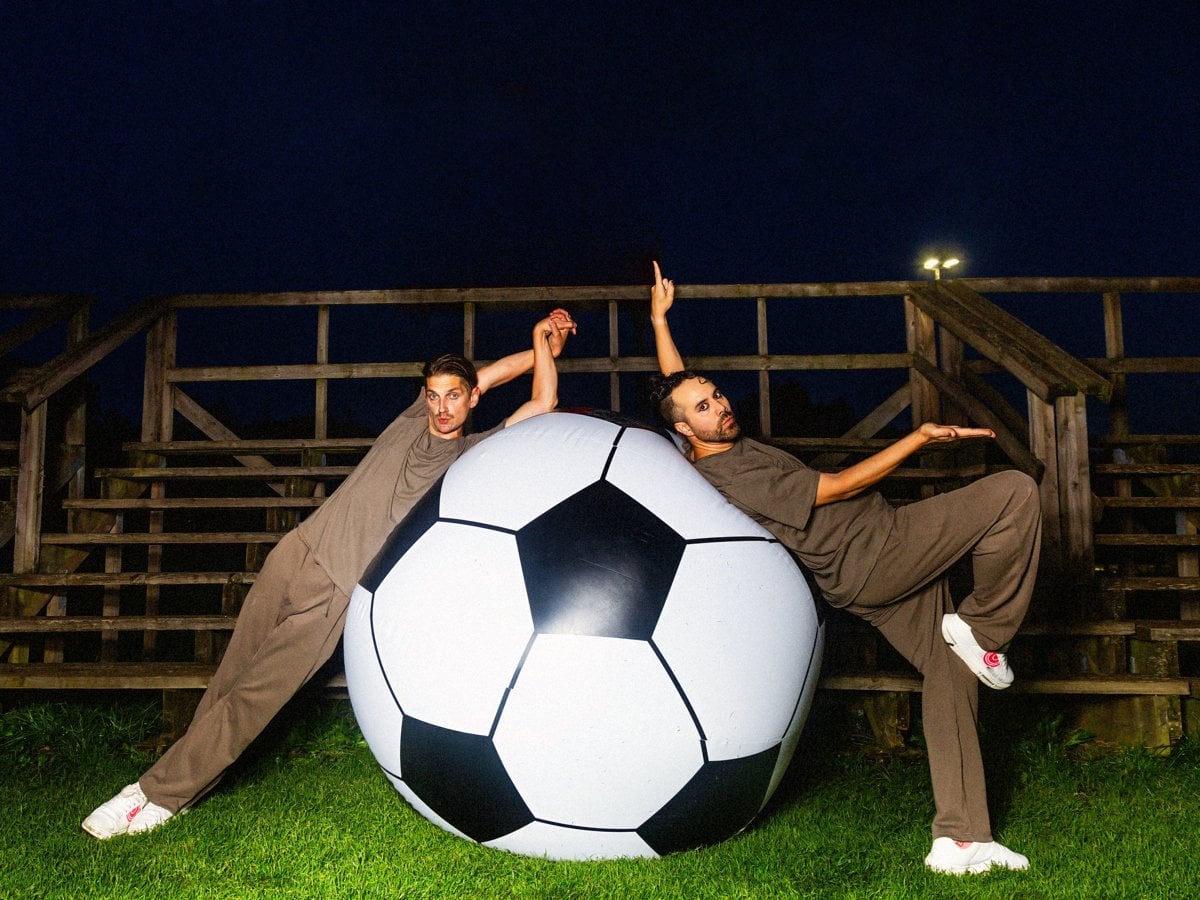 Två män i bruna träningsoveraller och vita sneakers lutar sig elegant mot en jättestor fotboll på en konstgräsplan framför en läktare på natten.