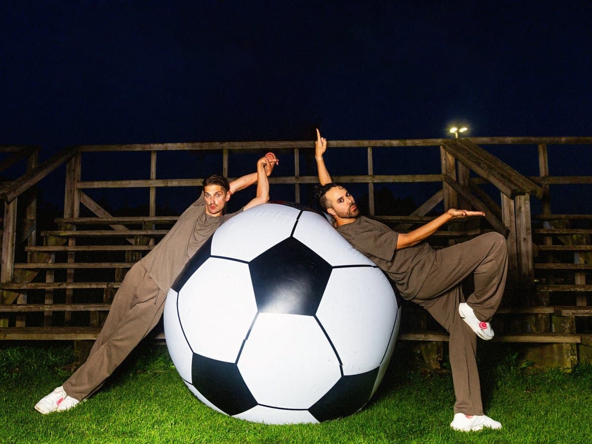 Två män i bruna träningsoveraller och vita sneakers lutar sig elegant mot en jättestor fotboll på en konstgräsplan framför en läktare på natten.