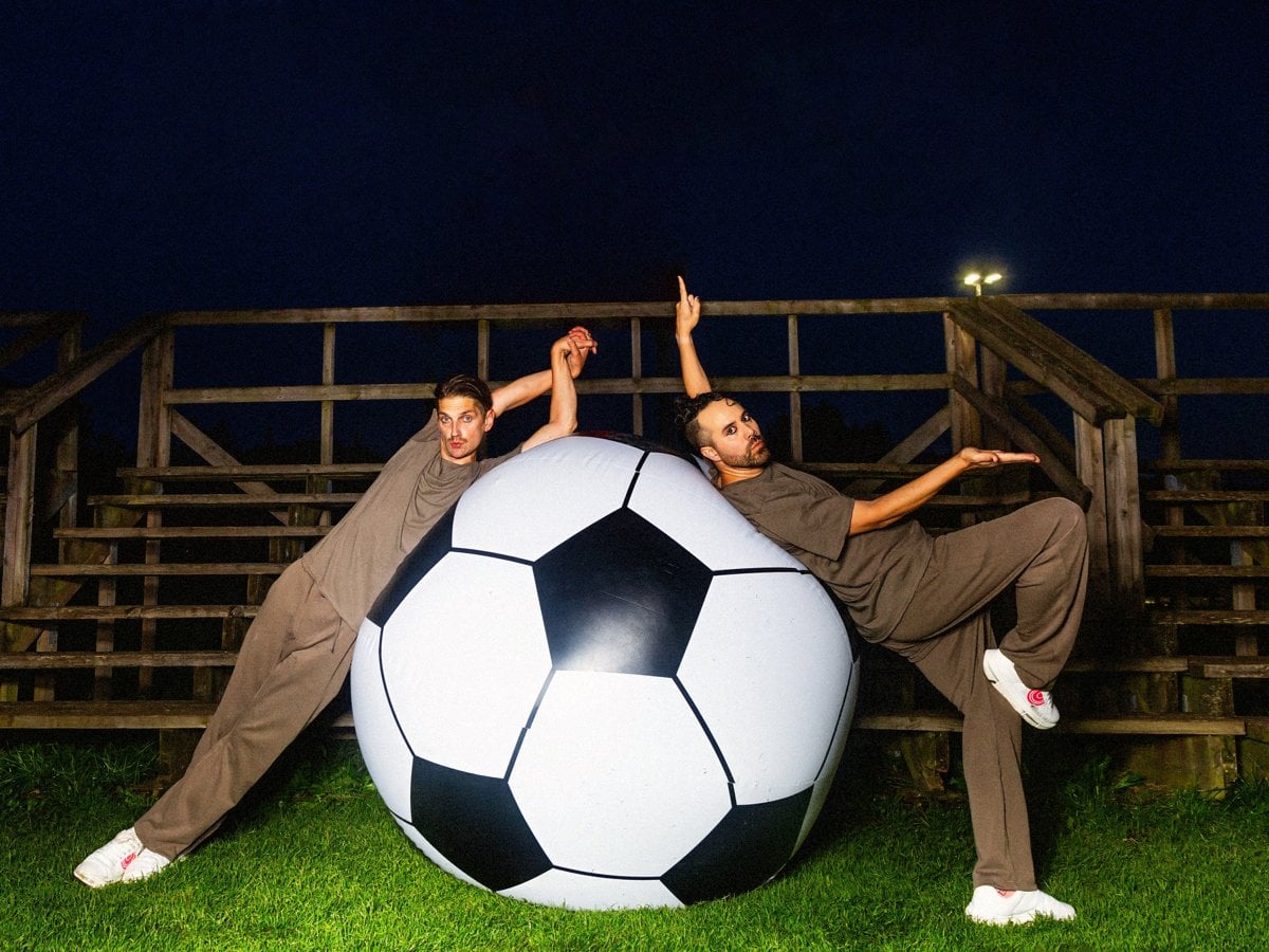 Två män i bruna träningsoveraller och vita sneakers lutar sig elegant mot en jättestor fotboll på en konstgräsplan framför en läktare på natten.