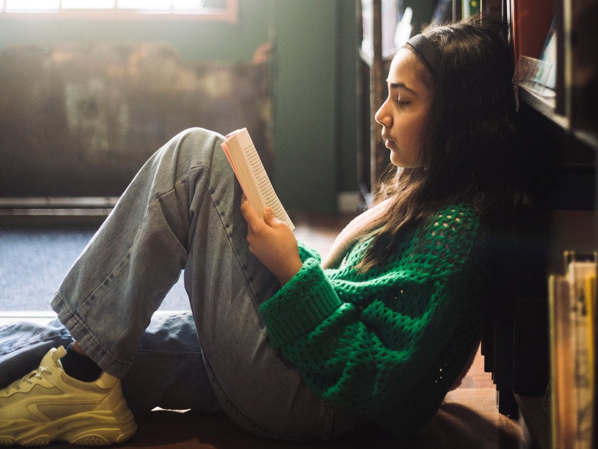 Flicka i tioårsåldern sitter lutad mot en bokhylla och läser en bok.