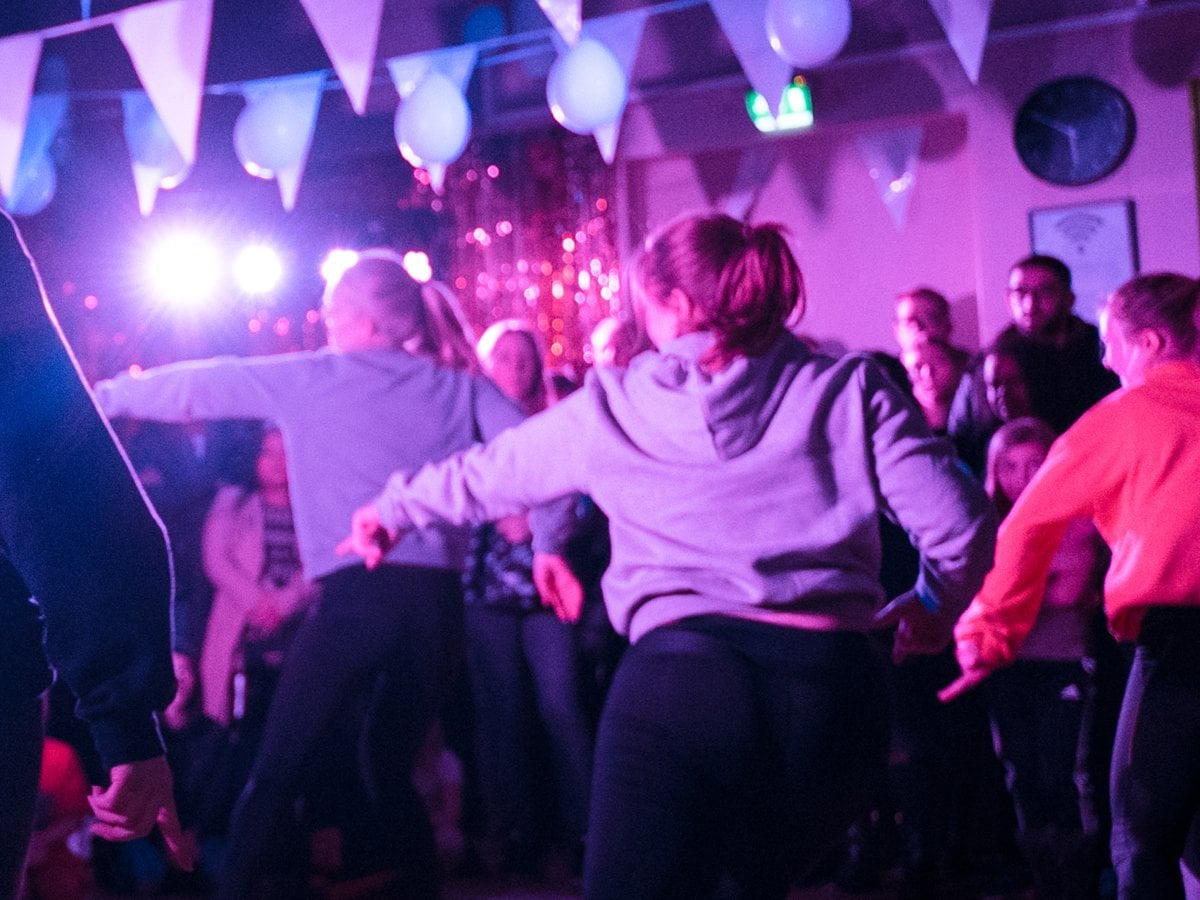 Fem dansare dansar en synkroniserad koreografi på ett dansgolv i en mörk lokal med flaggor, ballonger och festlig belysning i taket.