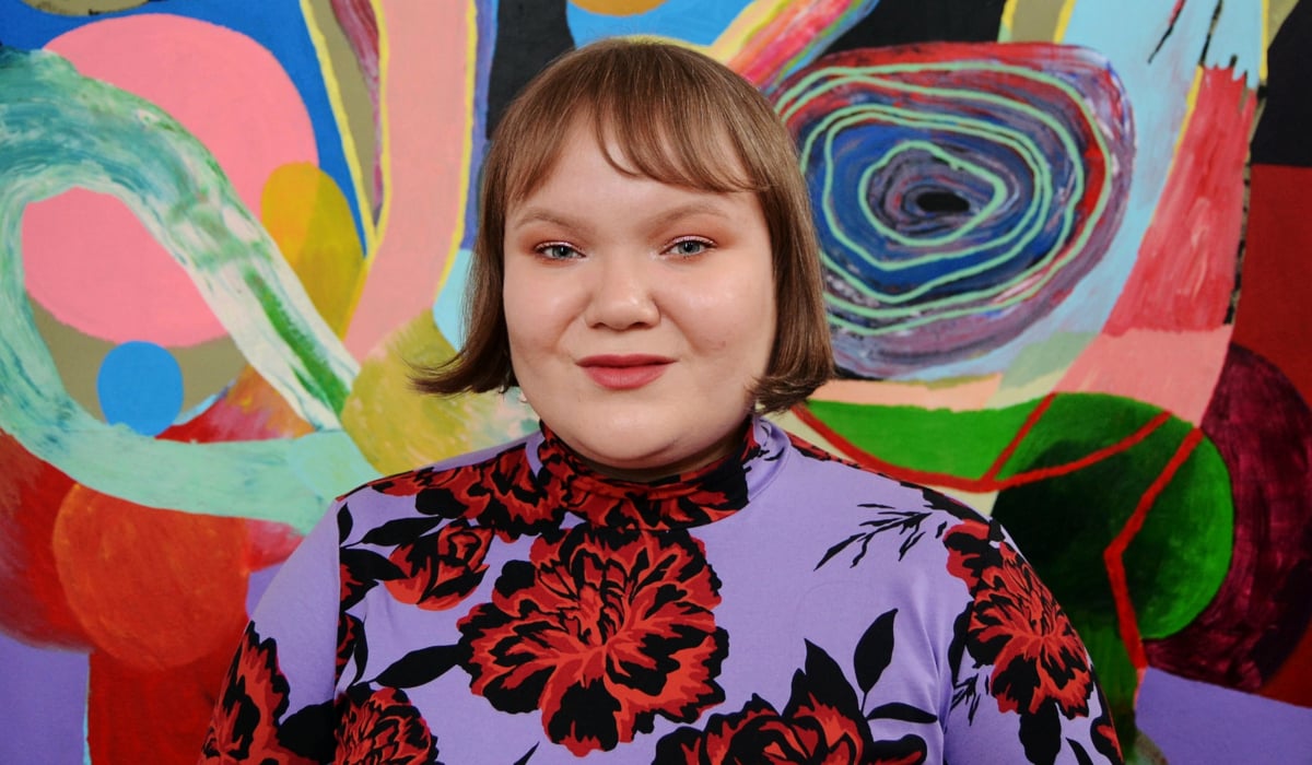 Linn framför en målning i abstrakt lekfullt mönster. Linn en mönstrad klänning med stora blommor.