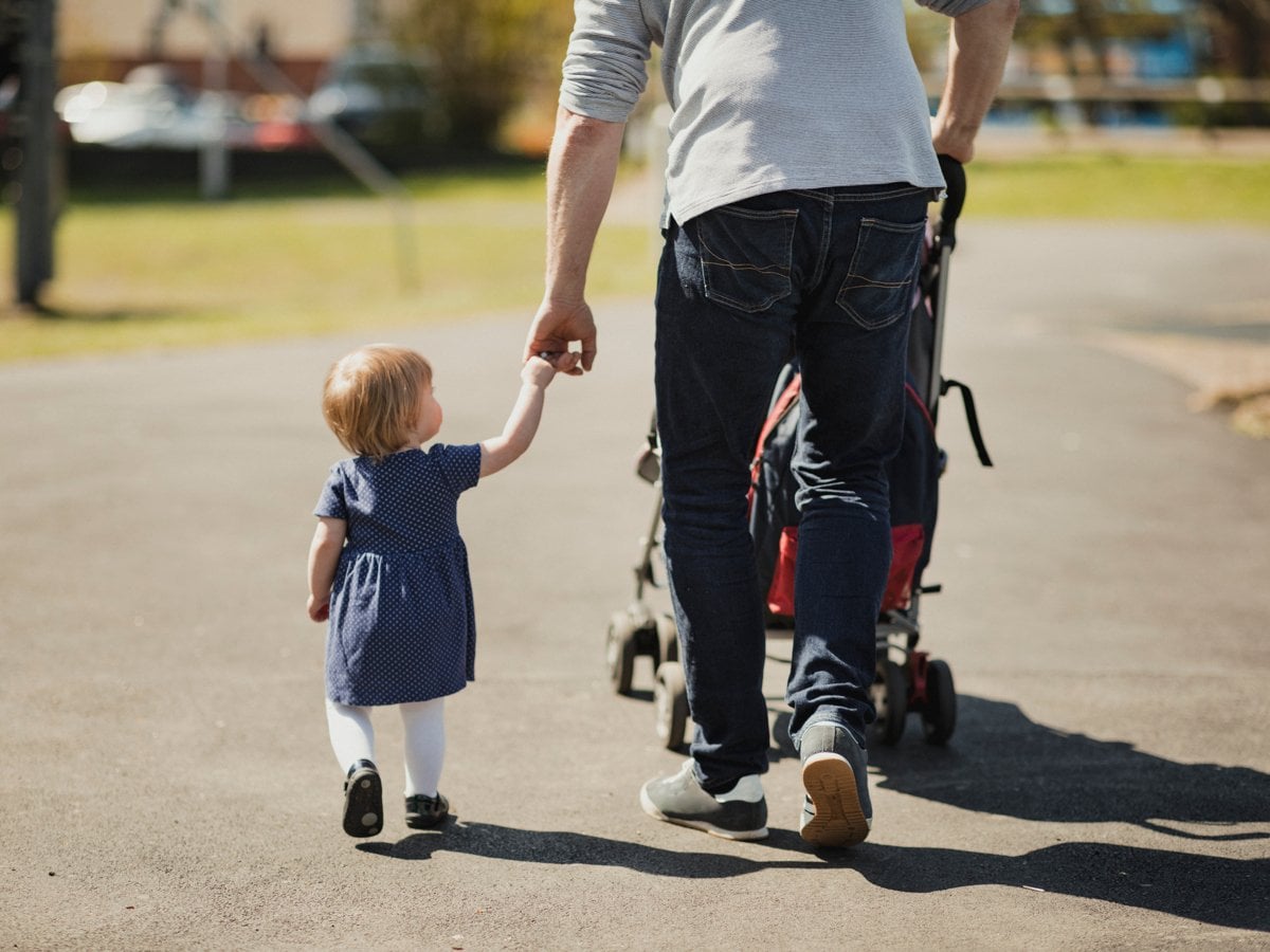 Liten flicka på promenad med sin pappa. Hon håller honom i handen och han i en barnvagn.