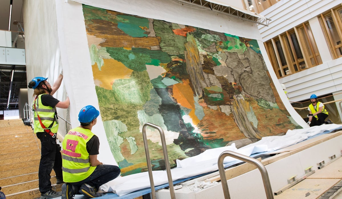 Tre personer i gula arbetsvästar justerar en stor målning på väg att hissas upp.