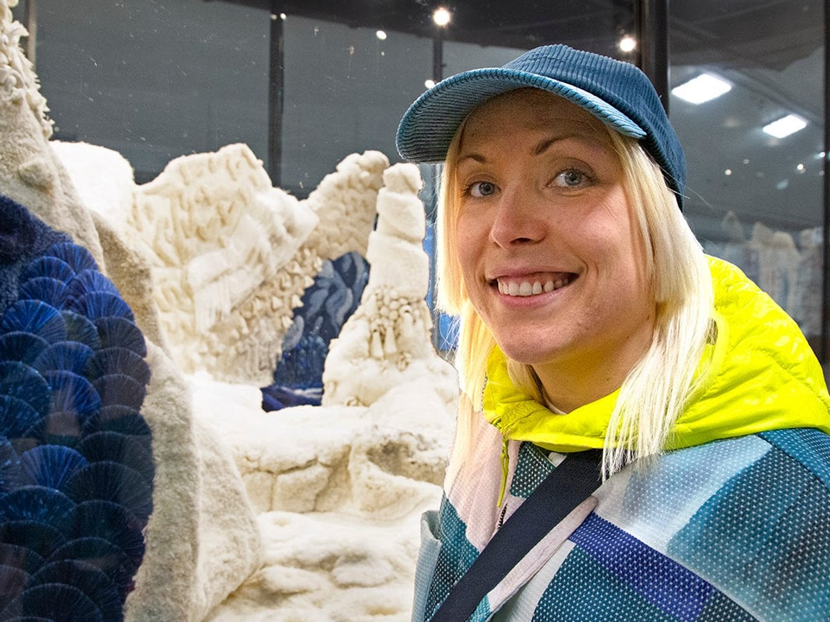 Sofia ler och tittar mot kamerian. Bakom henne glasmontern fylld med stora textilskulpturer, som mattor som växer upp mot taket.