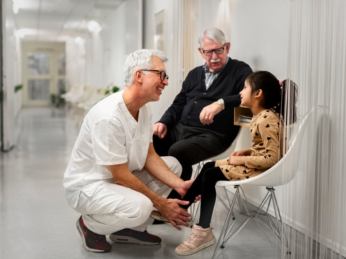 Manlig vårdanställd på huk i vårdkorridor, ler mot flicka på stol. Äldre man i bakgrunden.