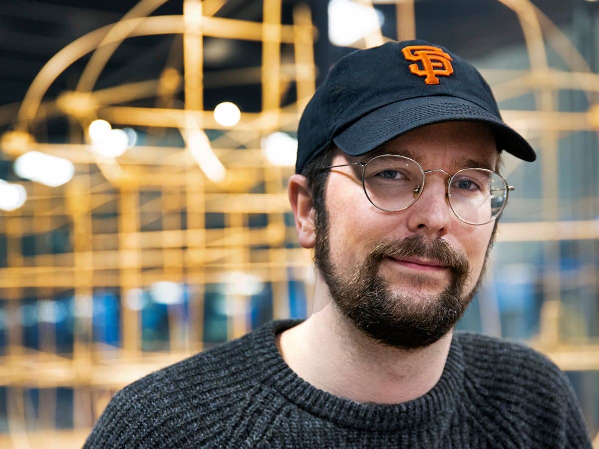 JEsper ler mot kamerian. Han har mörk keps och runda glasögonen och helskägg. I bakgrunden anas en stor träkonstruktion.