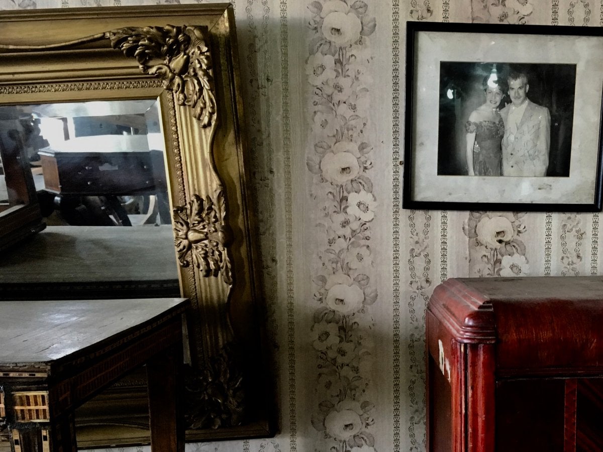 Stillbild från film. Dammigt bord och skåp under en spegel med guldram och ett fotografi som hänger snett på väggen.