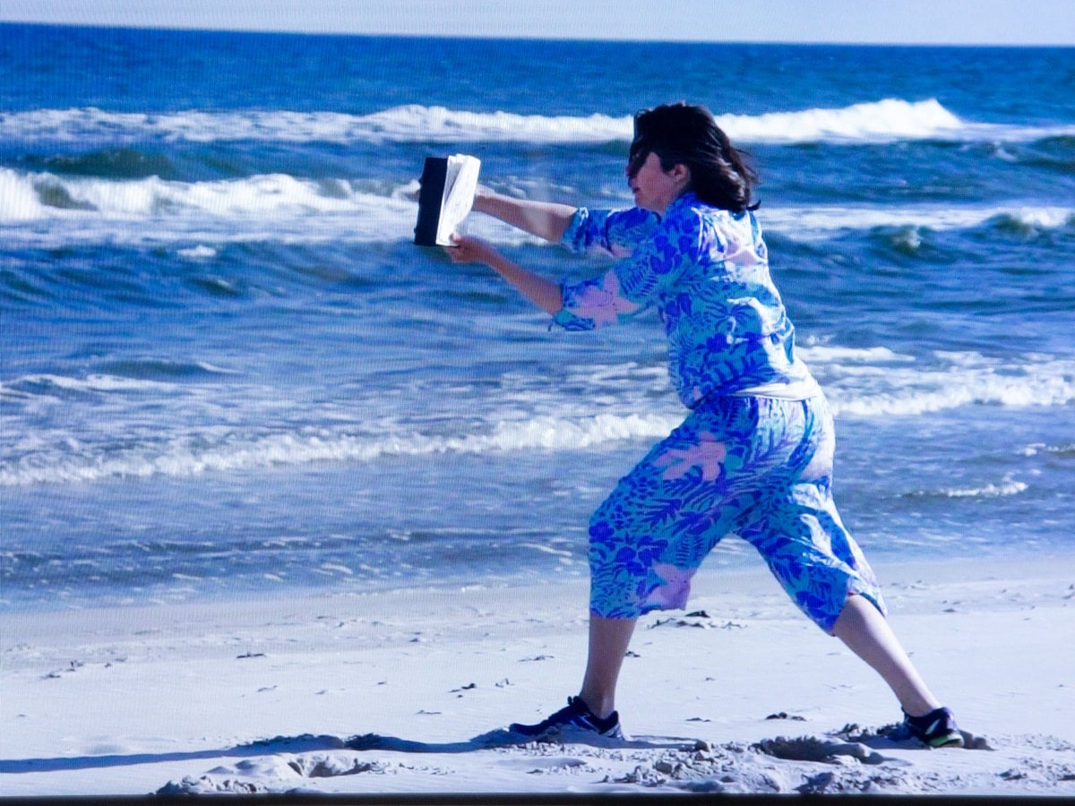 Anna står med en bok och läser i motvind på en sandstrand.