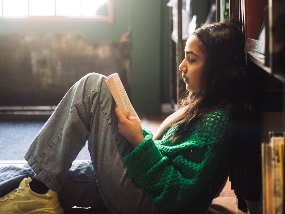 Flicka sitter lutad mot bokhylla och läser