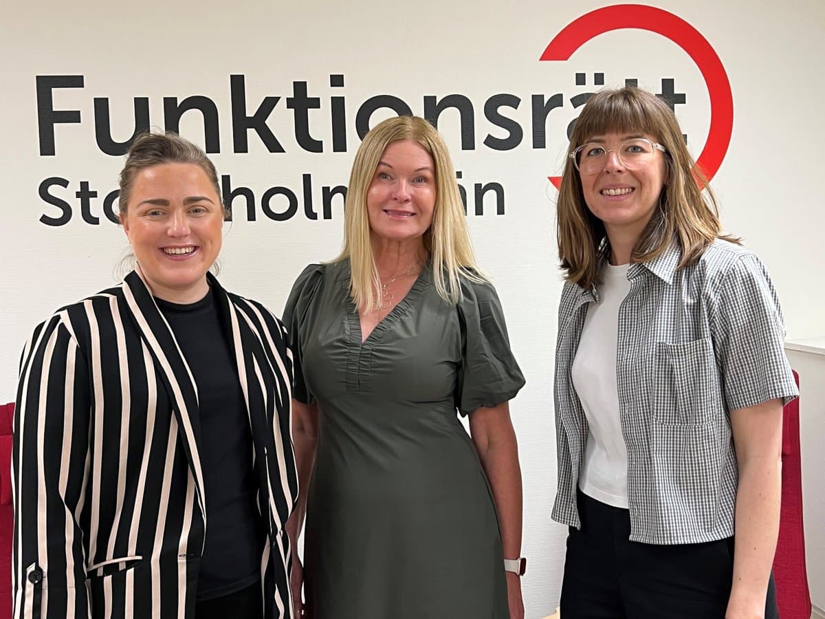 Tre kvinnor står bredvid varandra framför en skylt.