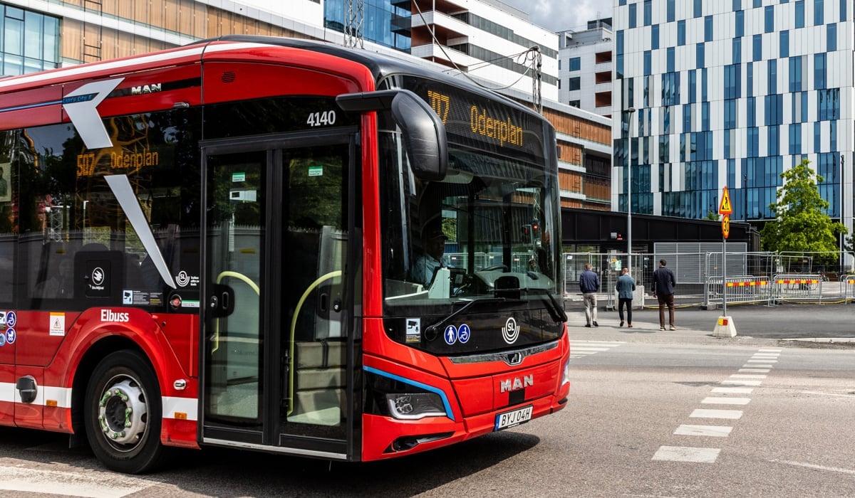 En röd buss kör längs en gata bredvid höga byggnader.