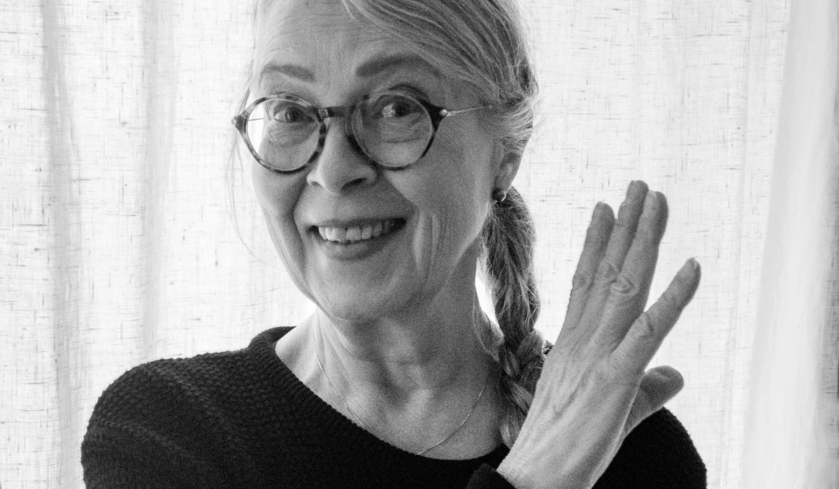 Ett svart-vitt fotografi av Kristina som ler mot kameran. Hon har lång fläta på ena sidan och håller en hand i luften.