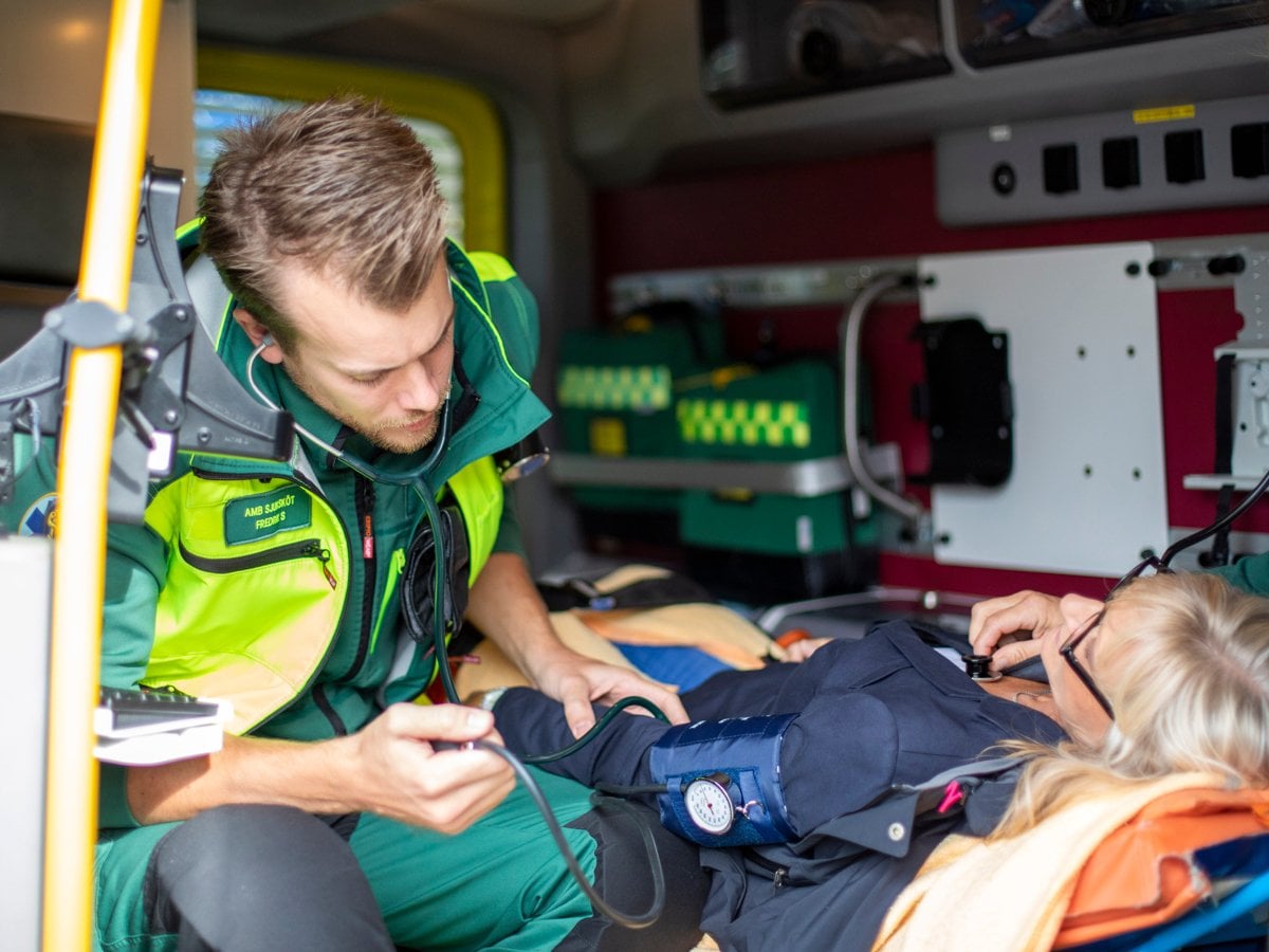 Medarbetare inom ambulanssjukvården i neongröna arbetskläder tar hand om en kvinnlig patient som ligger på en bår.