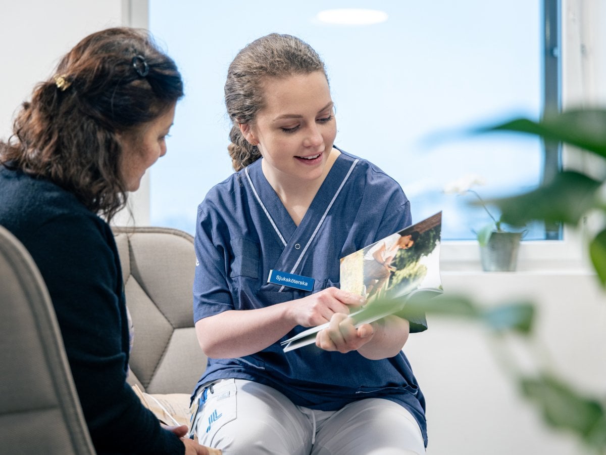 Två kvinnor sitter i en soffa och samtalar, en av kvinnorna är sjuksköterska och visar den andra kvinnan något i en broschyr.
