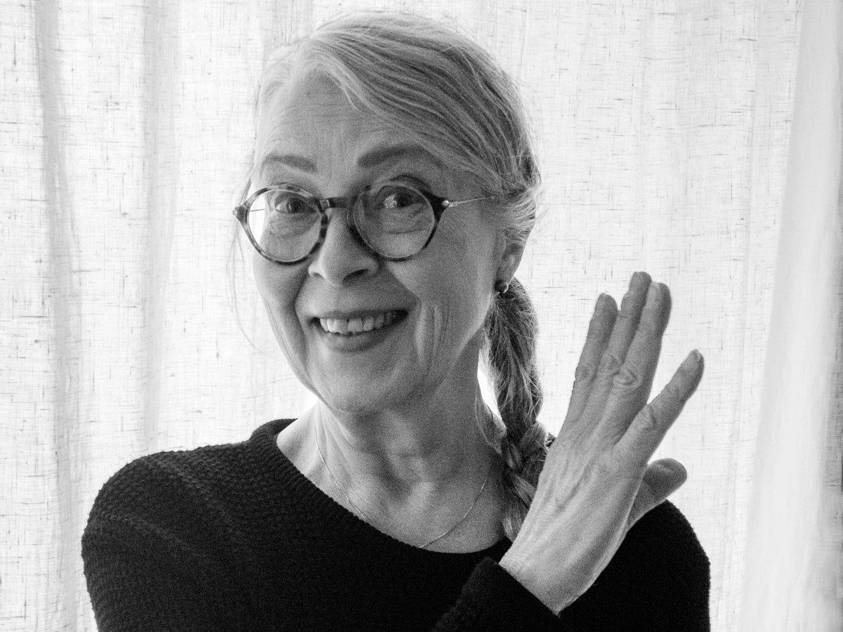 Ett svart-vitt fotografi av Kristina som ler mot kameran. Hon har lång fläta på ena sidan och håller en hand i luften.