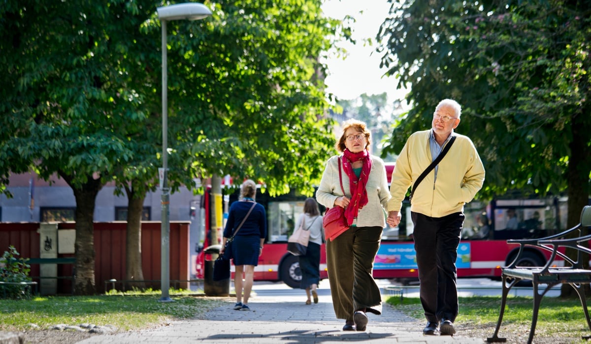 Äldre par promenerar i grön allé. SL-buss i bakgrunden.