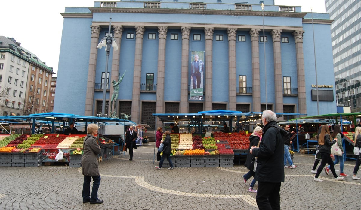 Människor går mellan Hötorgets fruktstånd. I bakgrunden det höga Konserthuset med sin pelare vid huvudingången.