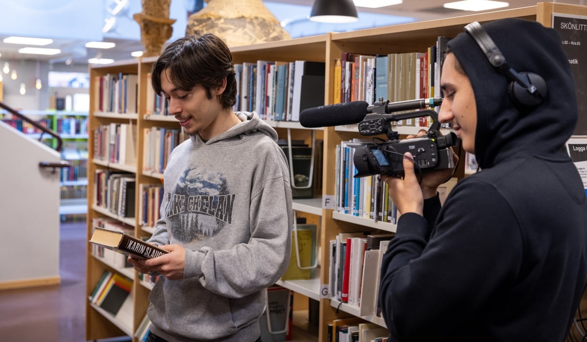 Två unga killar som gör film på ett bibliotek. En håller i en videokamera och har hörlurar på sig, en håller i en bok och blir filmad.