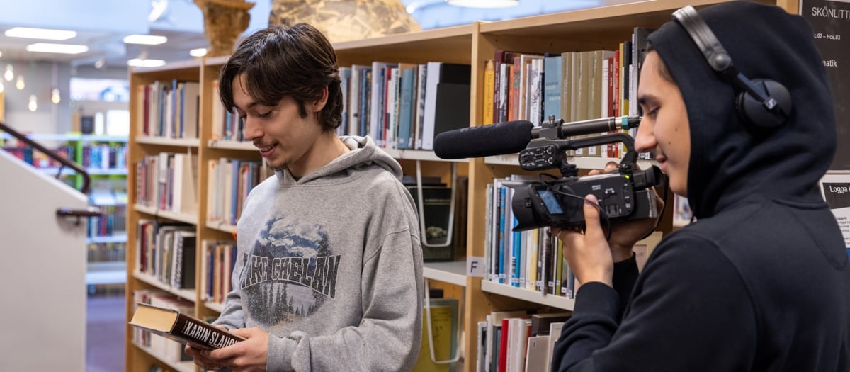 Två unga killar som gör film på ett bibliotek. En håller i en videokamera och har hörlurar på sig, en håller i en bok och blir filmad.