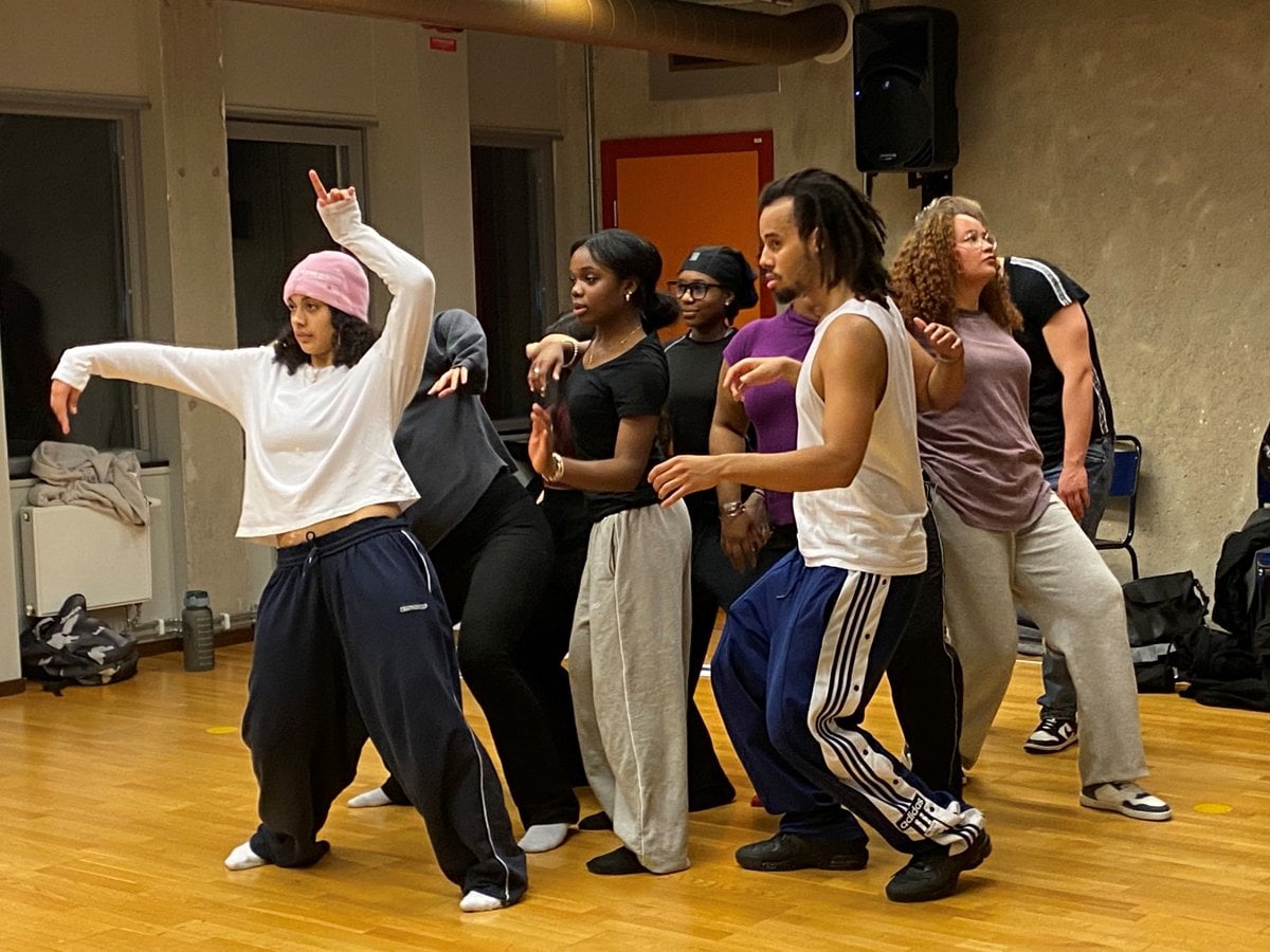 En grupp unga människor tränar dans  i en sal med trägolv.