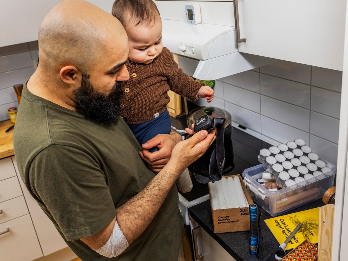 En man håller i en bebis i ett kök. Mannen tittar på utrustning för hemberedskap.