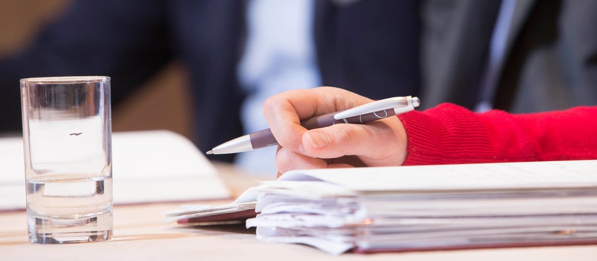 Närbild på en persons hand som håller i en penna. På bilden syns också en hög med papper och ett vattenglas.