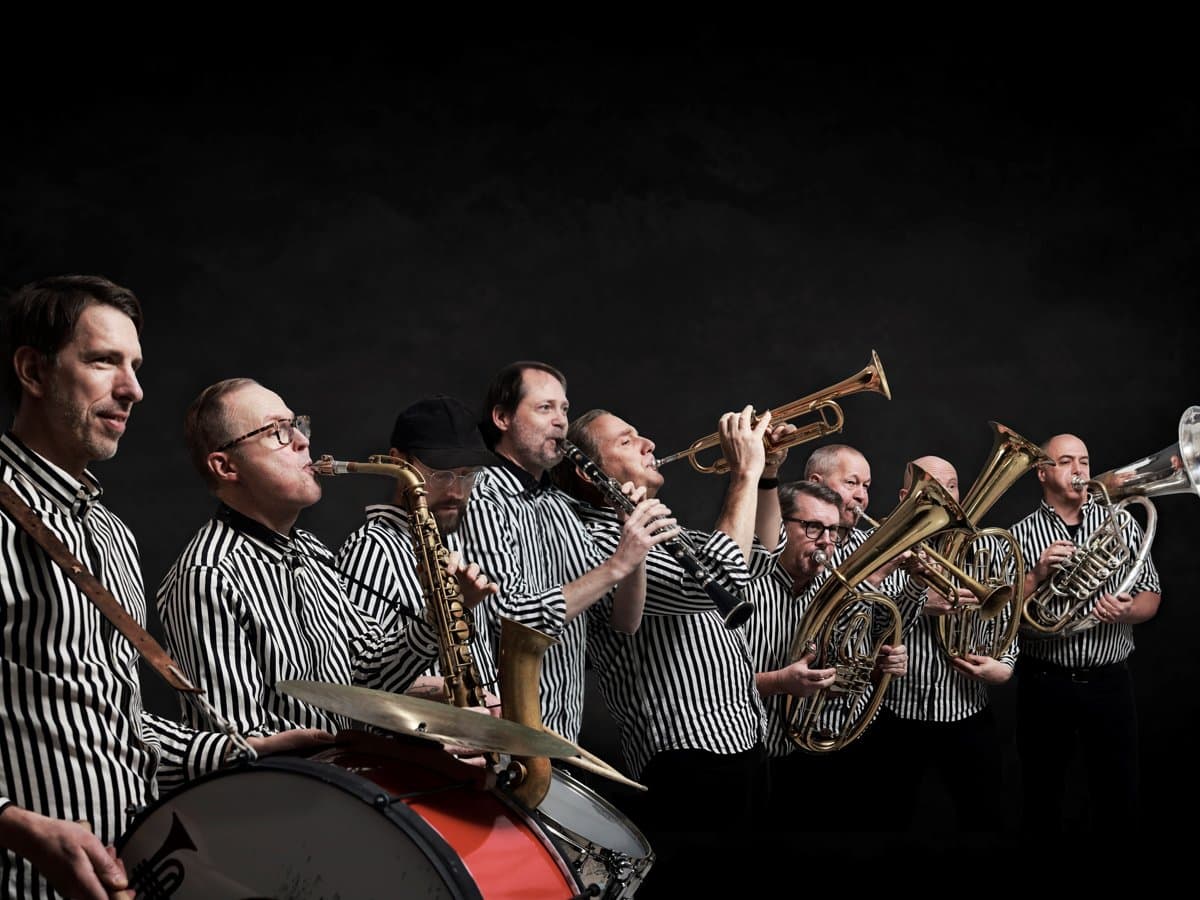 En grupp musiker med svartvitrandiga skjortor och instrument mot en svart vägg
