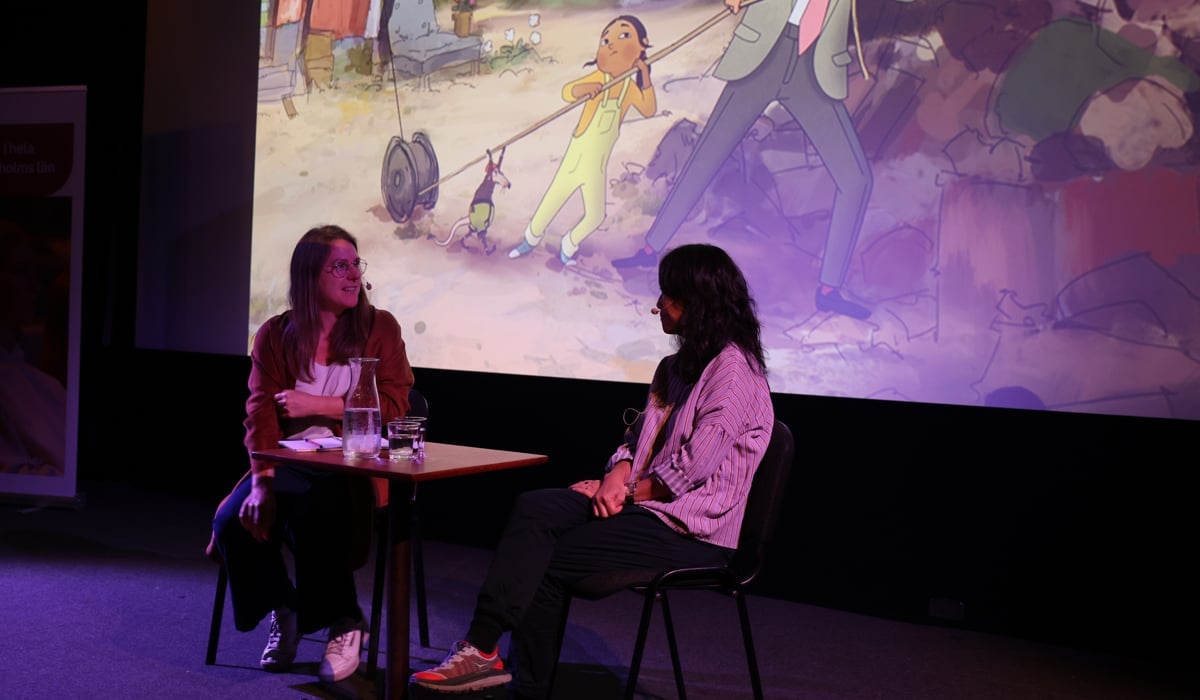 Två personer sitter och pratar framför bioduken i en biografsalong. På duken syns en bild från en animerad barnfilm.