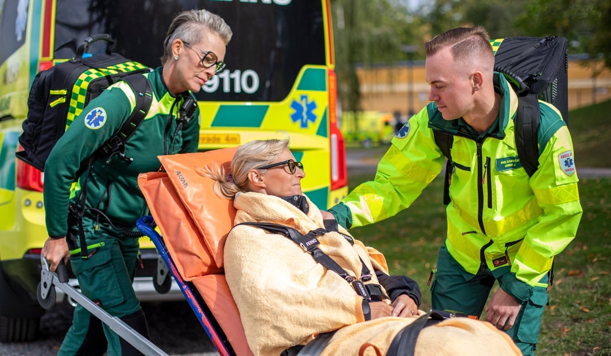 Två medarbetare inom ambulanssjukvården i neongröna arbetskläder tar hand om en kvinnlig patient som sitter på en bår.