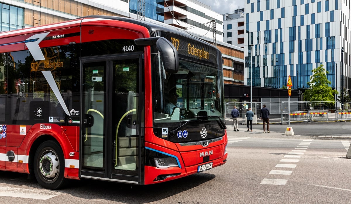En röd buss kör längs en gata bredvid höga byggnader.