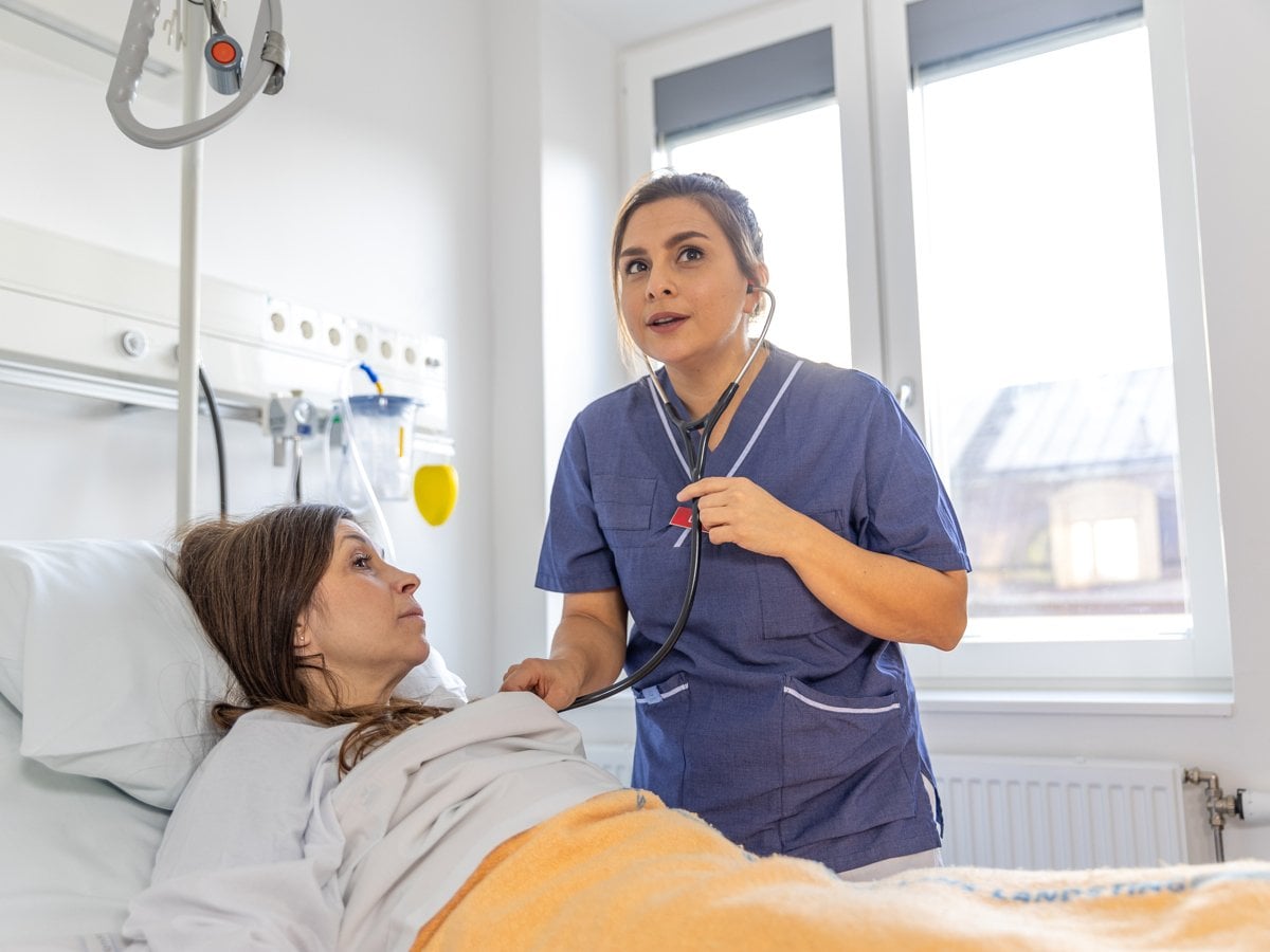 Kvinnlig läkare lyssnar med stetoskop på kvinnlig patient nedbäddad i sjukhussäng.