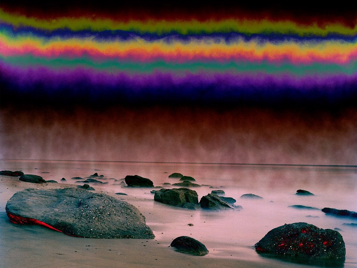 Flera stenar längst en strandkant. Vattnet är skymningsrosa och himlen är randig av olika ljusskimmer som en magisk regnbåge.